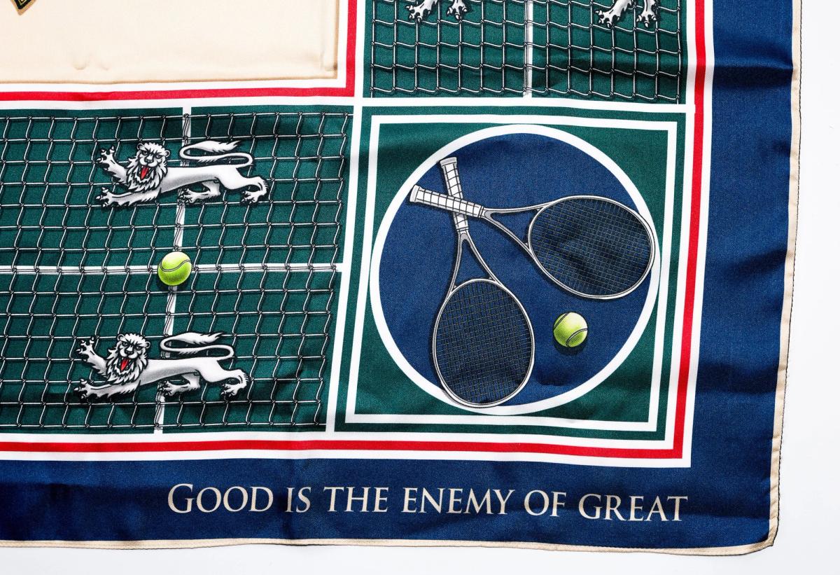 A corner of the scarf showing crossed tennis rackets and balls in a navy roundel over the green court, with the motto good is the enemy of great along the border.