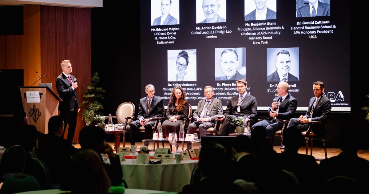A conference panel of six seated speakers on a stage beside a standing man at a lectern, with a large screen behind showing headshots and titles of the panellists.