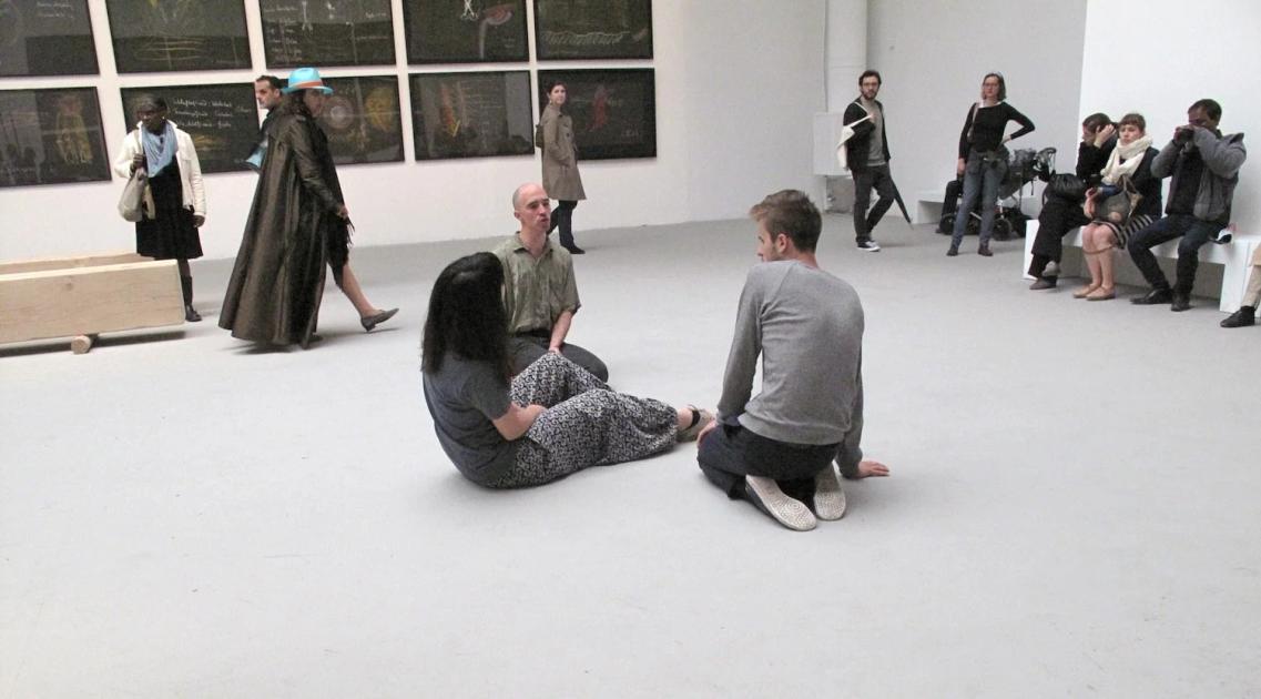 A white gallery space where visitors stand and sit on the floor while several people move among them, some with arms raised, framed works hung along the far wall.
