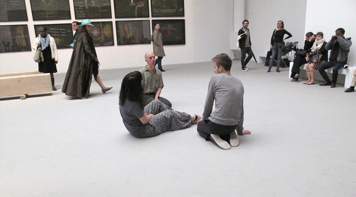 A white gallery space where visitors stand and sit on the floor while several people move among them, framed works hung along the far wall.