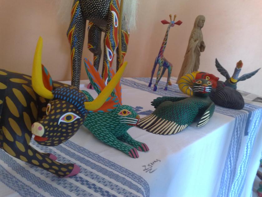 Brightly painted carved wooden animals arranged on a white cloth covered table, including a dark bull with yellow spots and curved horns, a green rabbit, a striped bird and a slender giraffe, with one unpainted standing figure in pale raw wood behind them.
