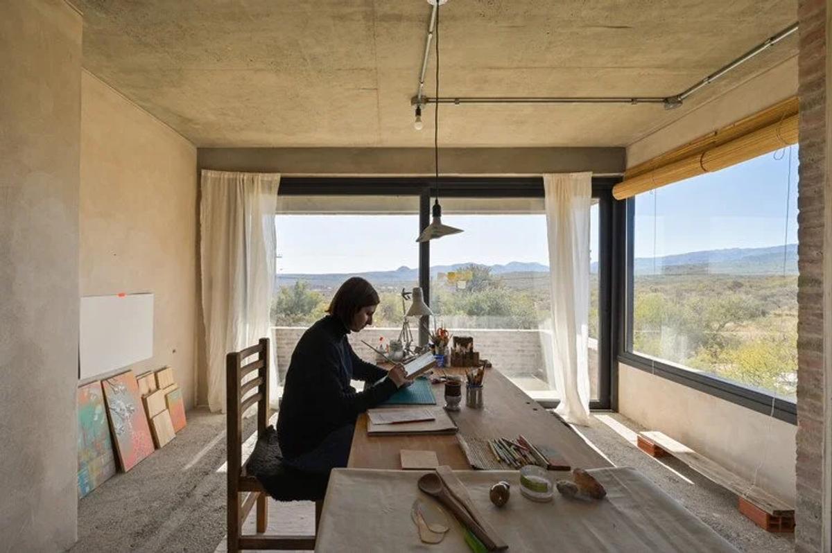 A person working at a long table in a bare concrete studio, a wide window looking out over open countryside and unframed canvases stacked against the wall.