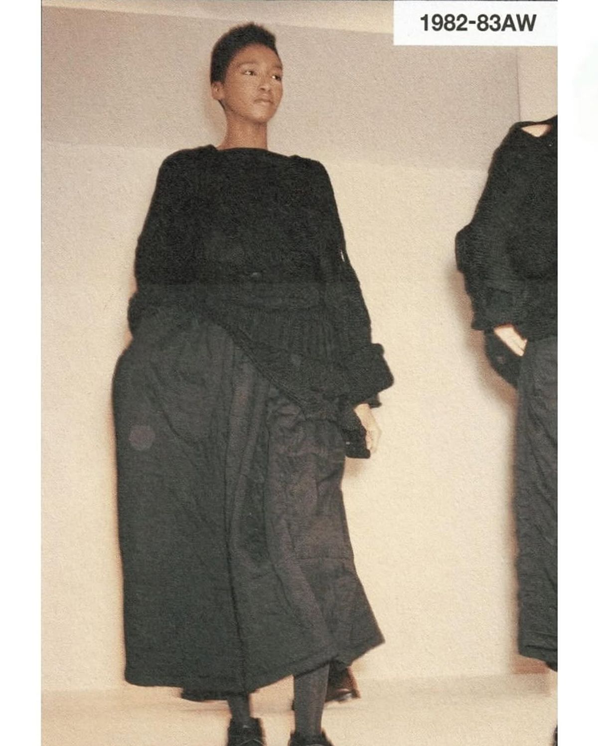 A photograph labelled 1982 to 83 autumn winter, a model standing in a long dark layered wrap garment with a second figure behind.