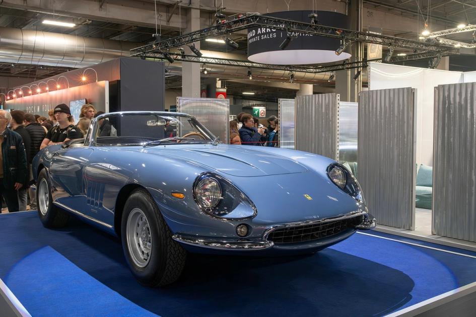 A pale blue 1967 Ferrari 275 GTB/4 NART Spider on a blue display plinth under show lighting, with an exhibition banner overhead and visitors in the background.