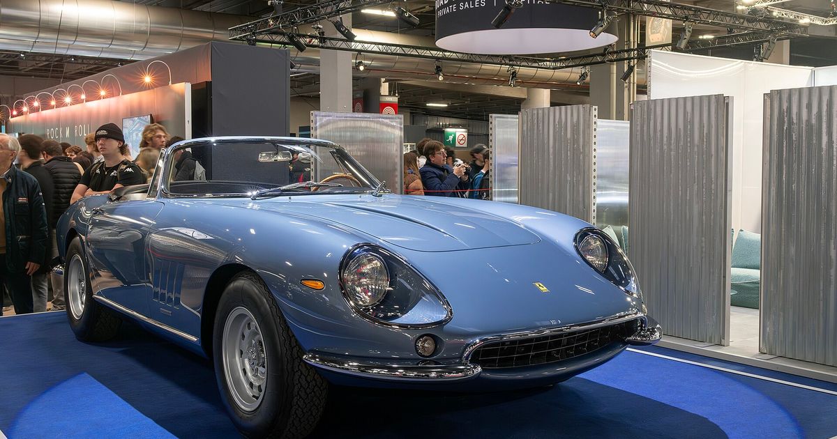 A pale blue 1967 Ferrari 275 GTB/4 NART Spider on a blue display plinth under show lighting, with an exhibition banner overhead and visitors in the background.