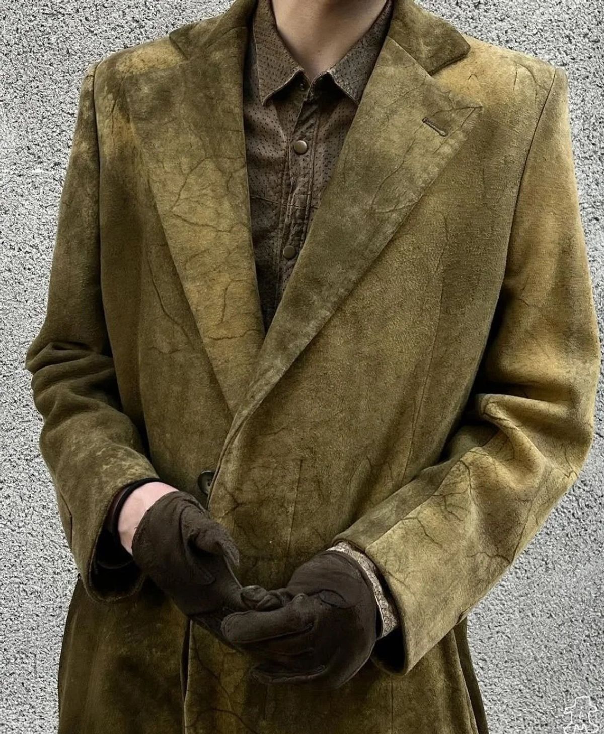 A person photographed from the neck down in a long olive suede coat over a matching shirt, hands clasped in dark gloves against a grey wall.