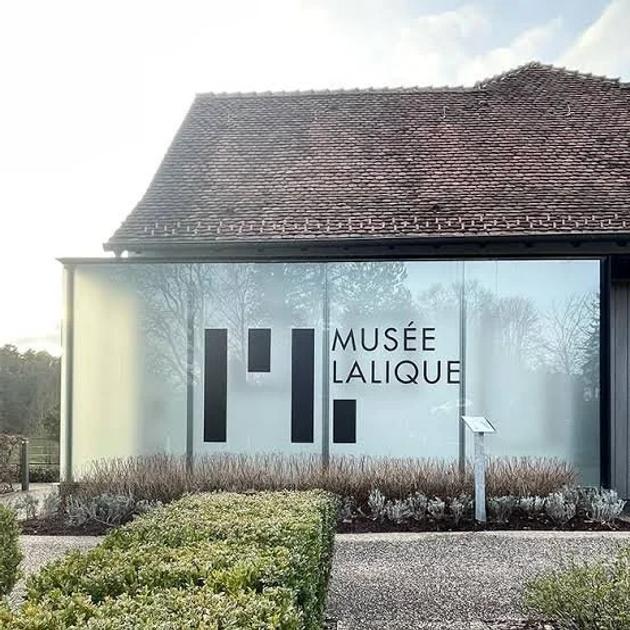 A low museum building with a tiled roof and full height glass walls, Musee Lalique lettering across the facade and clipped hedges in front.