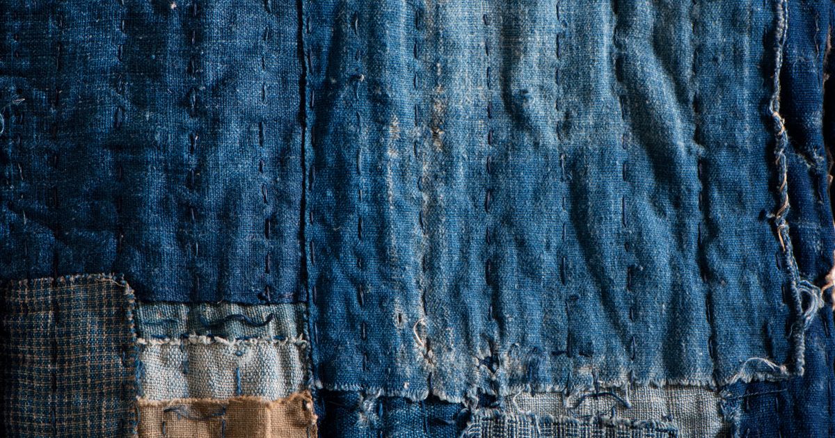 Close view of indigo cloth built from overlapping patches, crossed by long rows of pale running stitch, edges frayed.