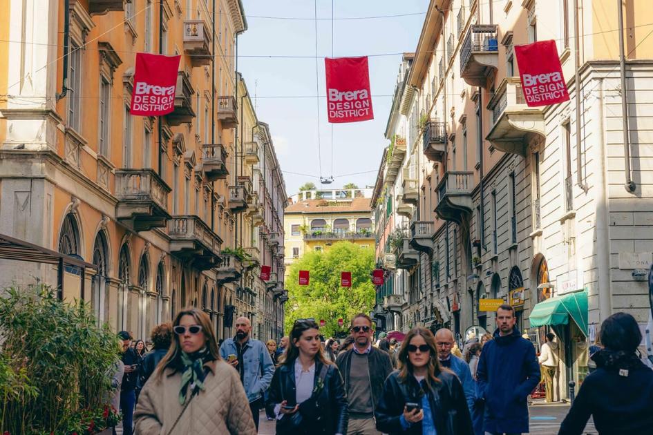 A crowded Milan street during design week, red Brera Design District banners strung between the buildings and pedestrians filling the road.