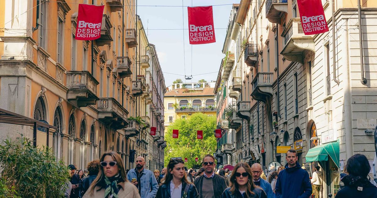 A crowded Milan street during design week, red Brera Design District banners strung between the buildings and pedestrians filling the road.