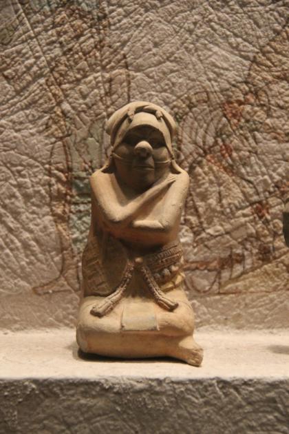 A small Maya clay figurine of a seated human figure in an elaborate headdress, photographed against black.