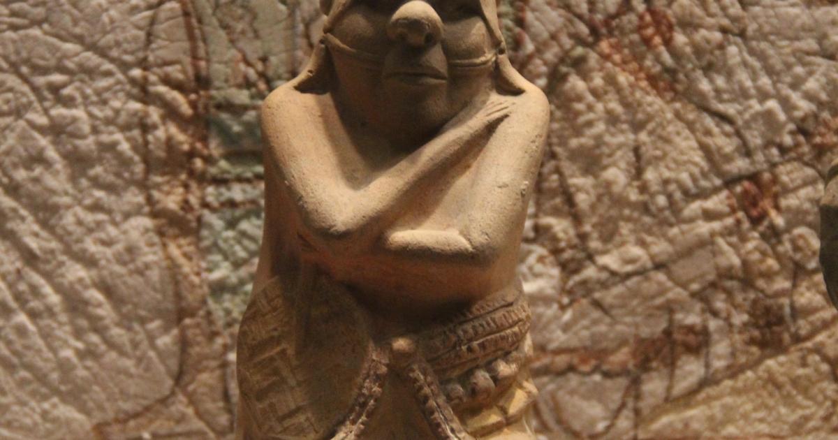 A small Maya clay figurine of a seated human figure in an elaborate headdress, photographed against black.