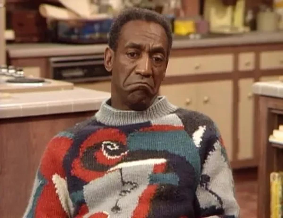 A man standing in a domestic kitchen wearing a bold patterned knitted sweater in red, cream and blue, looking to one side.