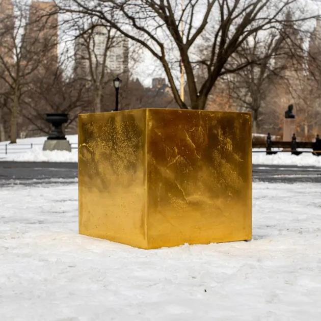 A large gold cube standing on snow in a city park, bare trees and railings behind it and buildings visible in the distance.