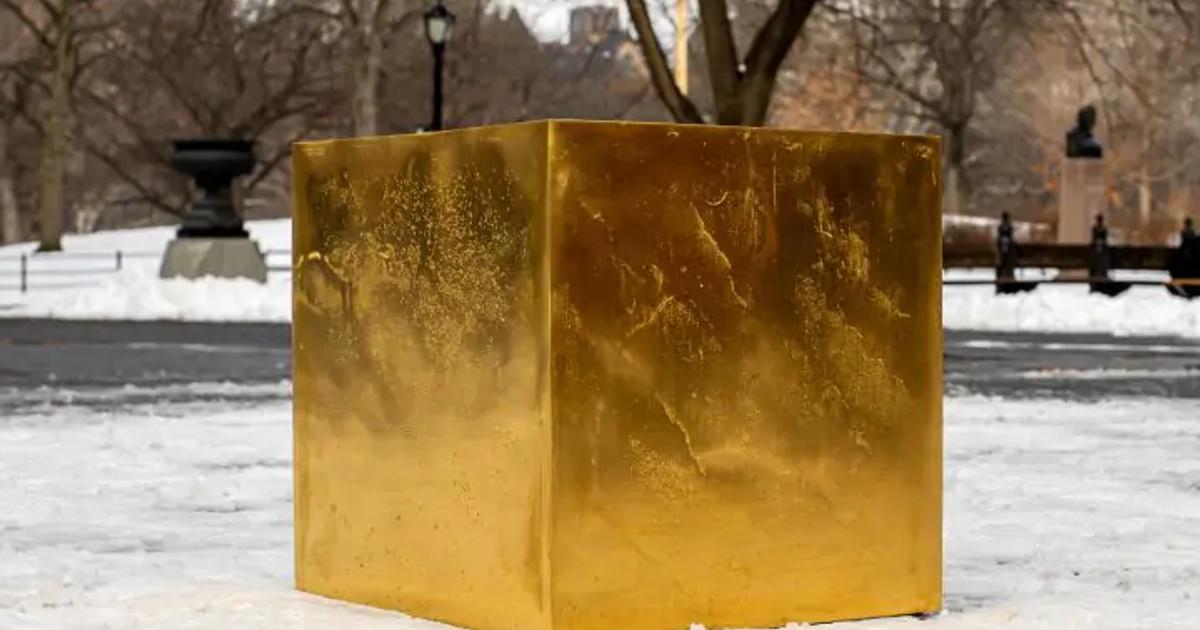 A large gold cube standing on snow in a city park, bare trees and railings behind it and buildings visible in the distance.