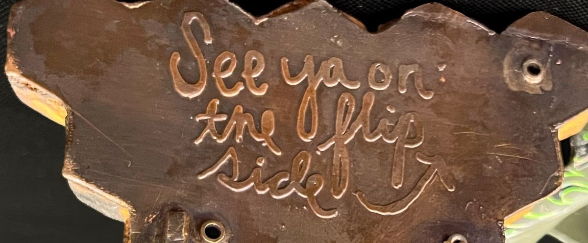 A close detail of hand cut cursive lettering pressed into copper, reading: See ya on the flip side.