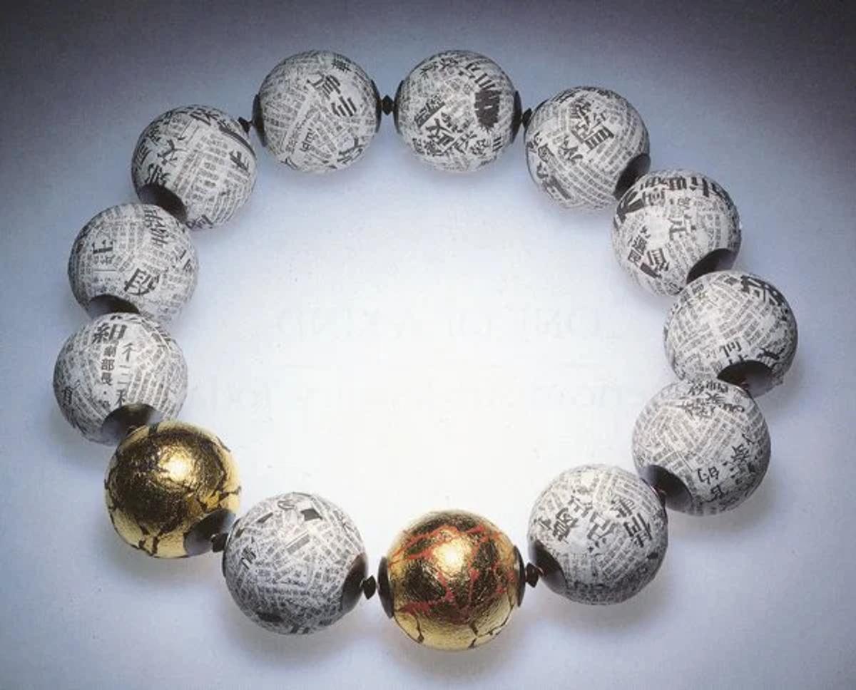 A necklace of large spherical beads covered in newsprint under clear resin, with three polished gold spheres set among them, arranged in a circle.