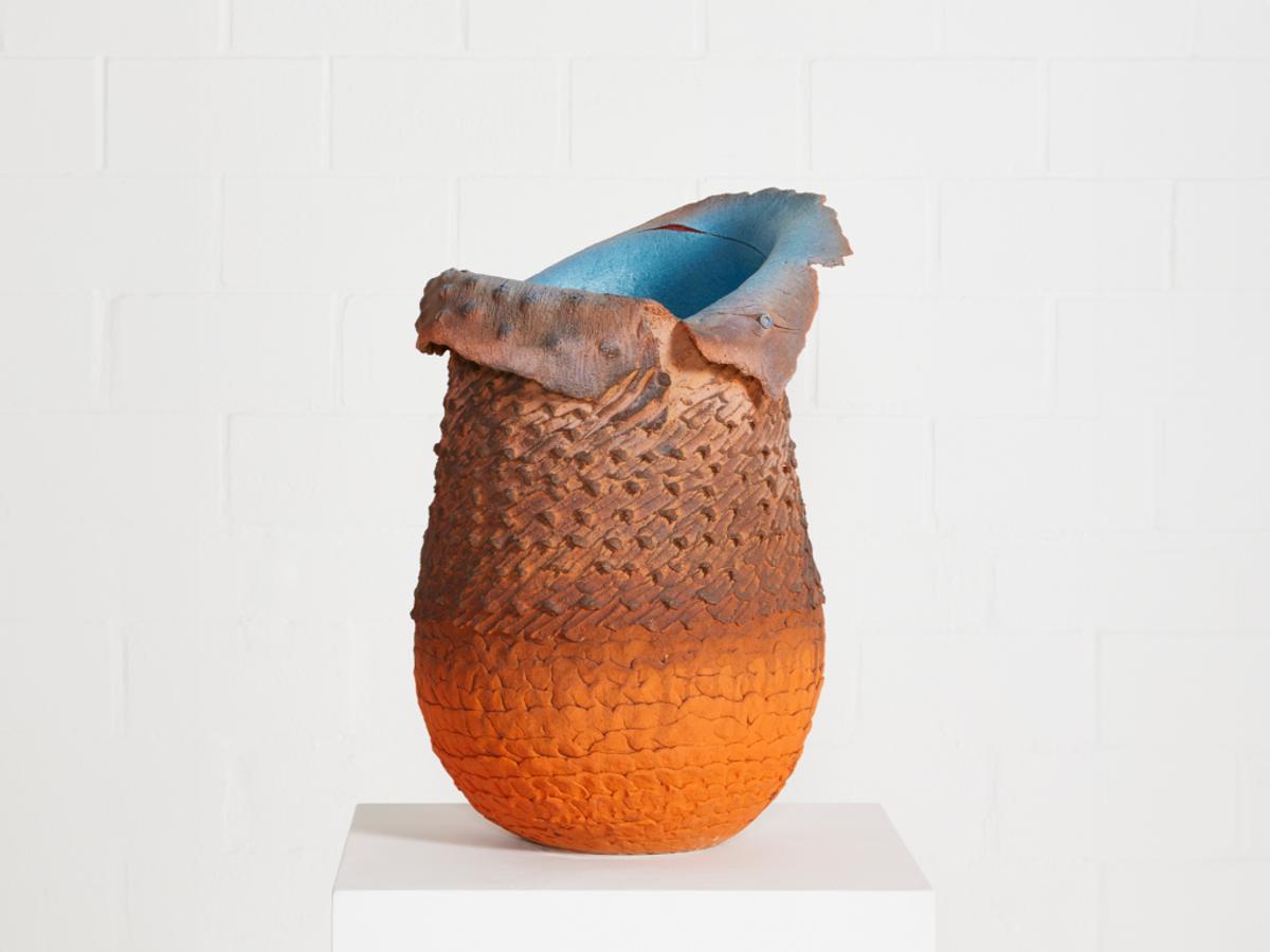 A tall ceramic vessel with a densely textured surface glazed from rust brown into bright orange, its rim torn open and pale blue inside, on a white plinth.