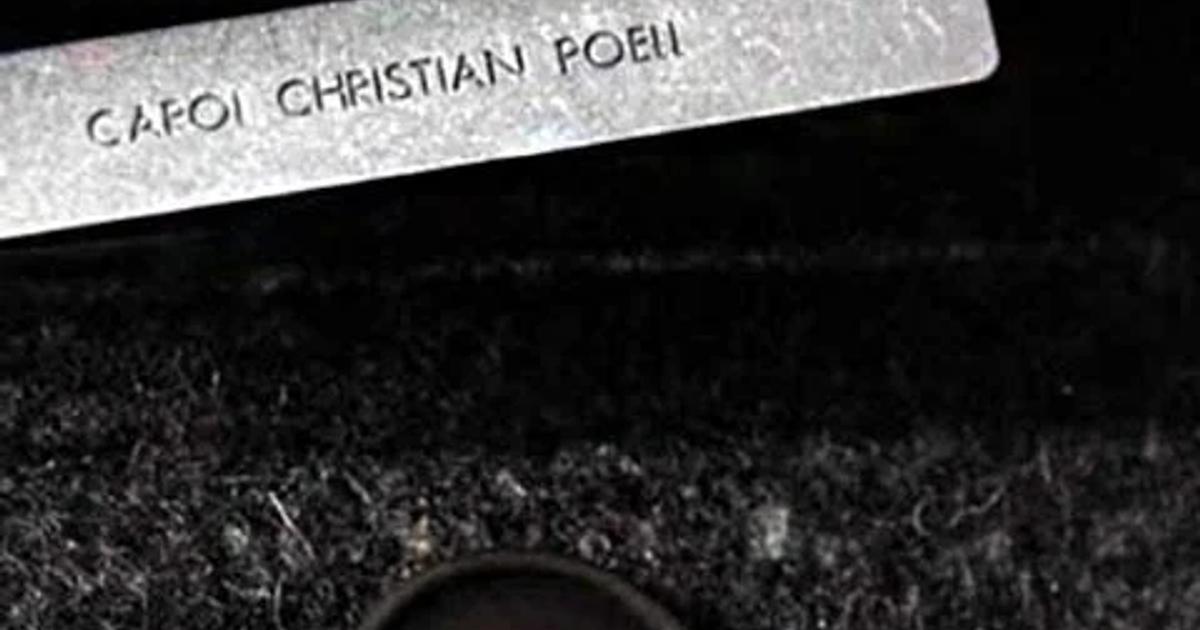 A close view of a stamped metal garment label sewn into heavy dark wool, with a dark four hole button below it.
