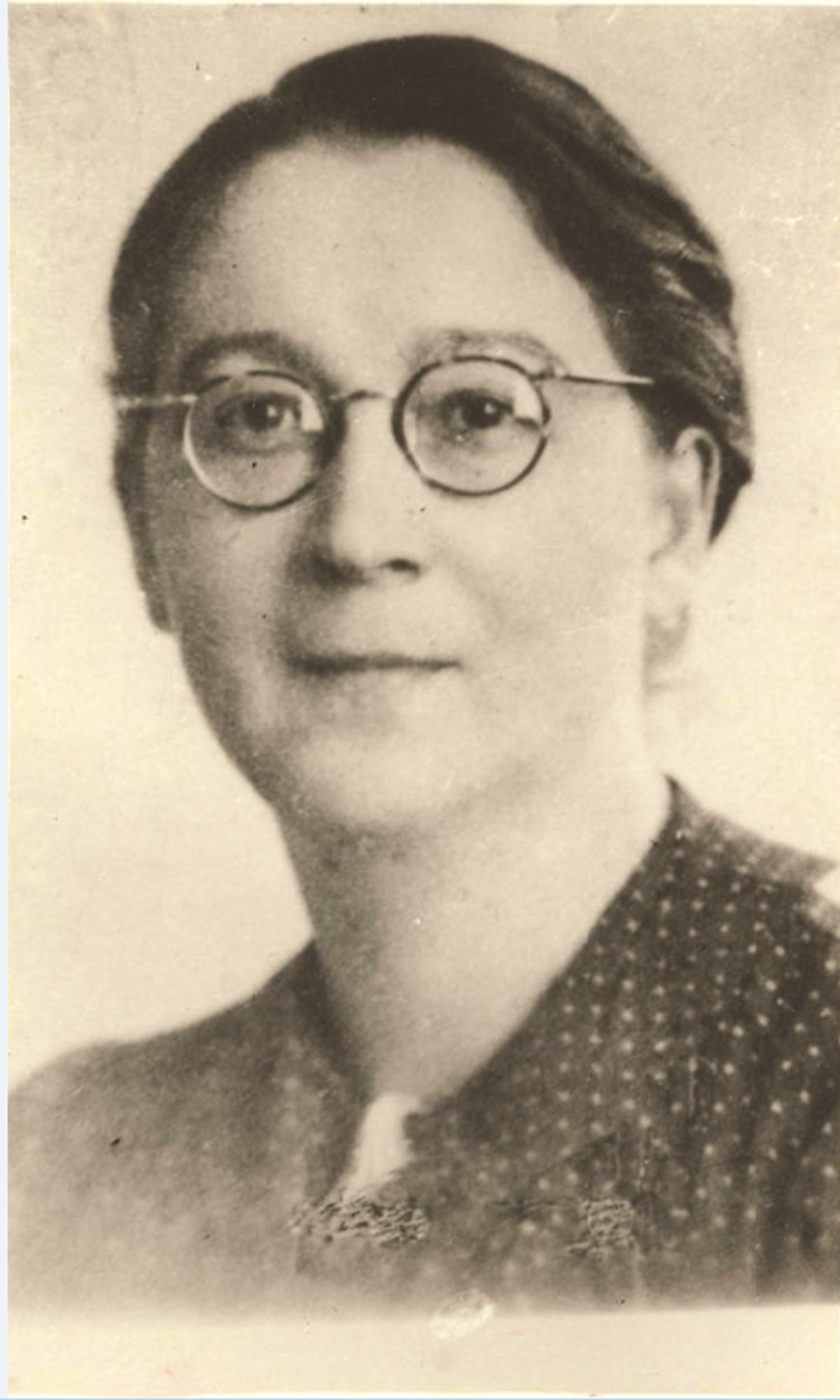 A black and white portrait photograph of a woman in round spectacles and a patterned dark dress, hair pinned back.