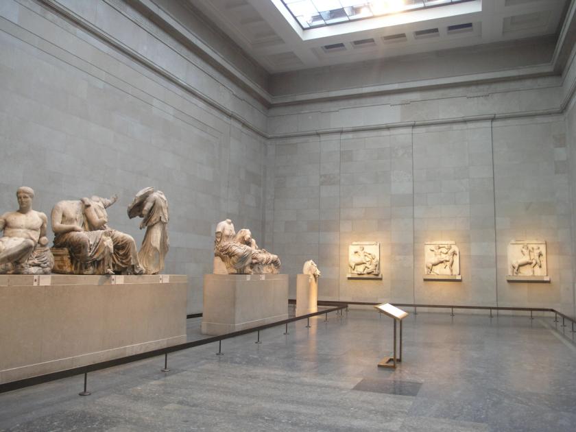 A long top-lit stone gallery with carved marble figures on low plinths along the left wall and relief panels mounted along the right.