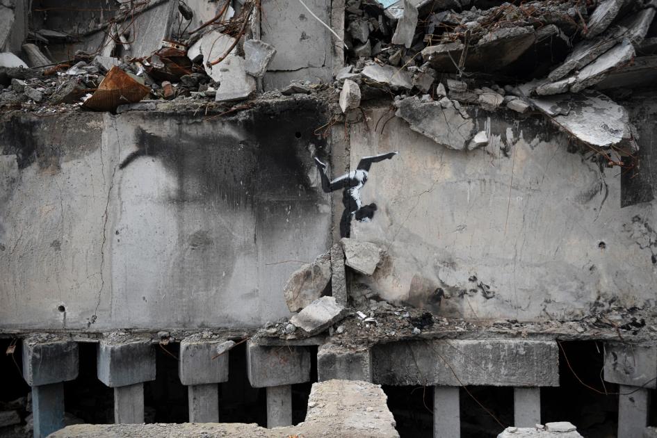 A bombed concrete building with its upper floors collapsed, and on the intact lower wall a stencil of a figure balanced on the rubble.