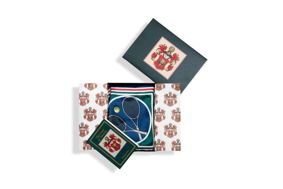 A boxed presentation set fanned out, a cloth folder and cards printed with a red and gold lion crest beside a sheet showing crossed tennis rackets.