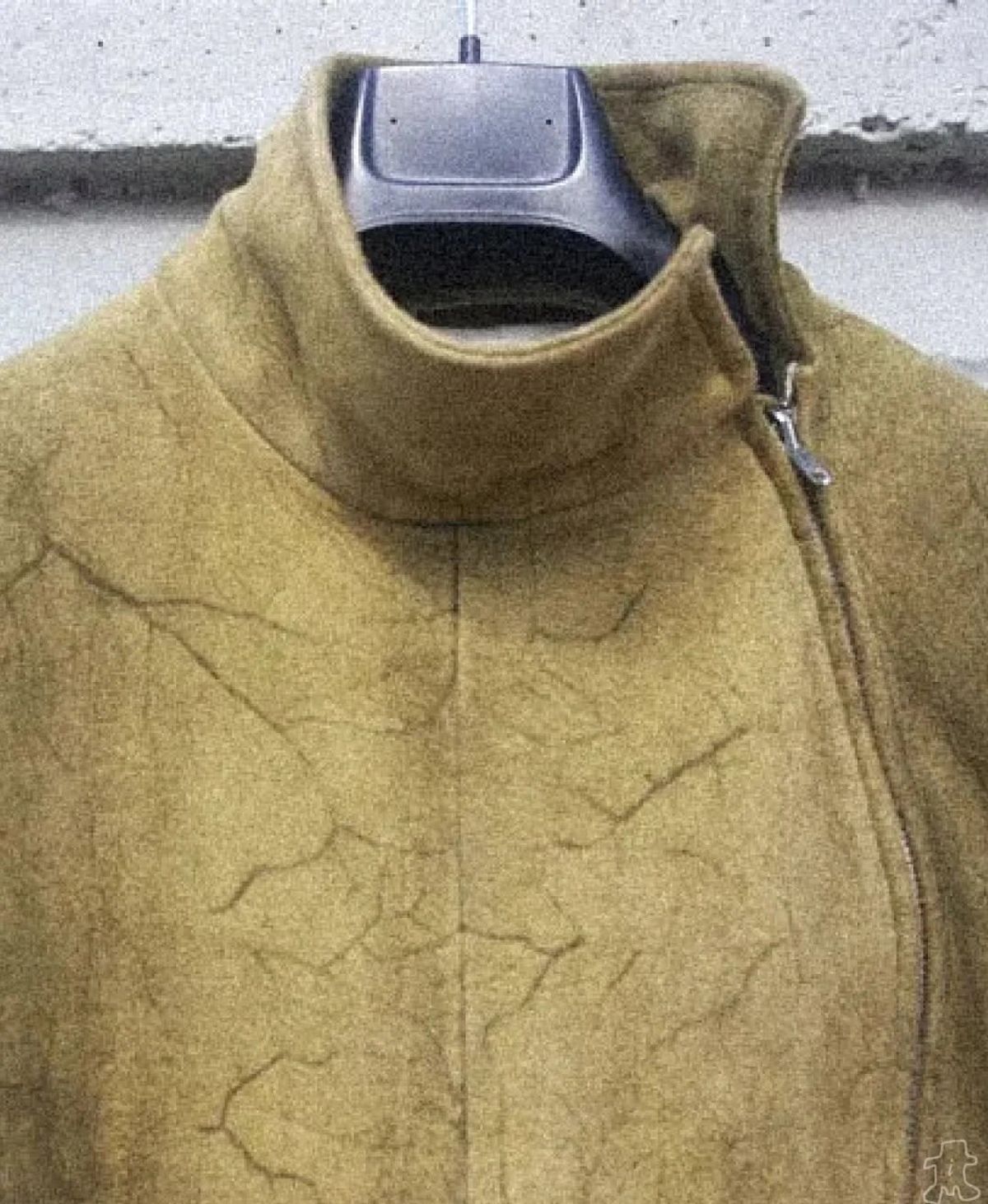 The upper half of an olive coated jacket on a hanger, its high funnel collar and chest showing a fine cracked surface like dried mud.