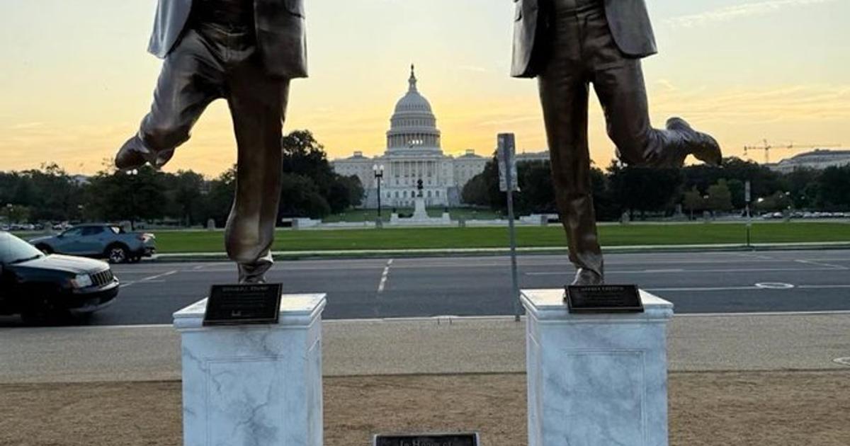 Two bronze statues of men in suits on white marble plinths, holding hands and kicking one leg up, with the United States Capitol dome behind them at dusk and a small plaque between the plinths.