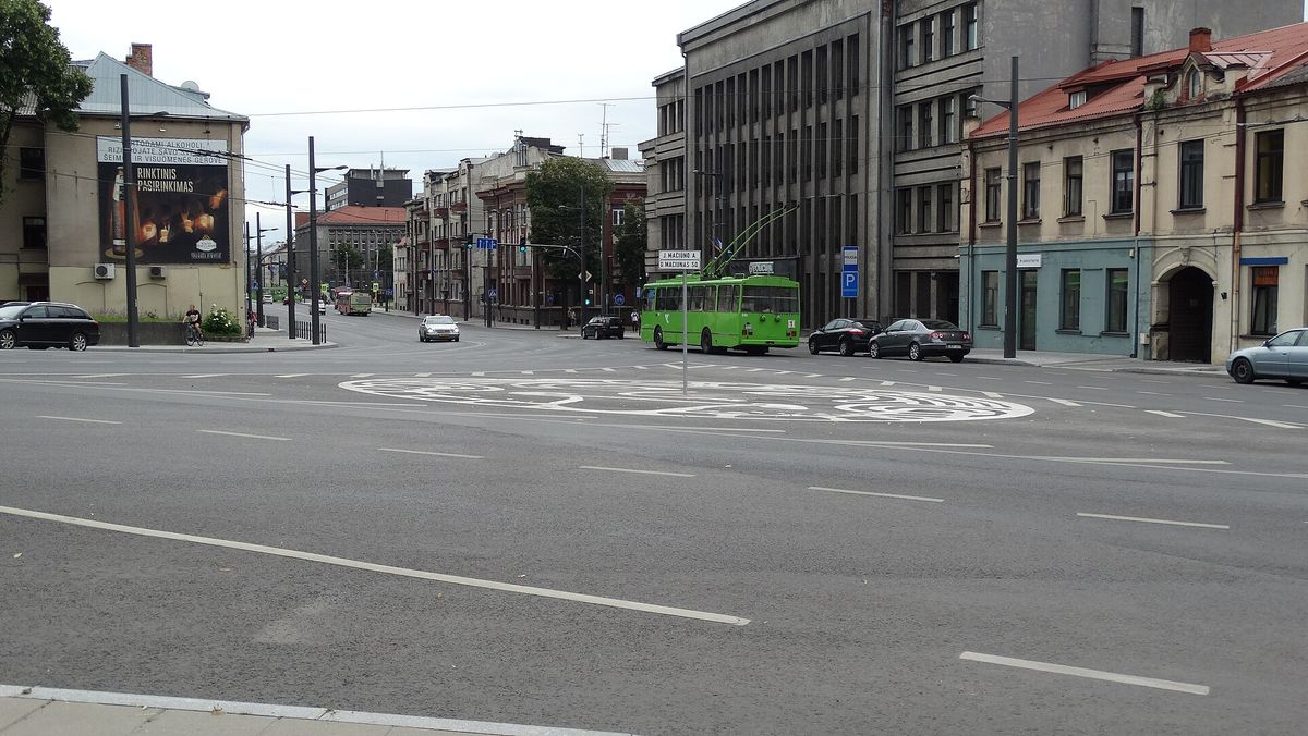 A wide road junction in Kaunas with a large white maze-like design painted across the asphalt, a street sign reading G. Maciunas Sq., and a trolleybus crossing the far side.