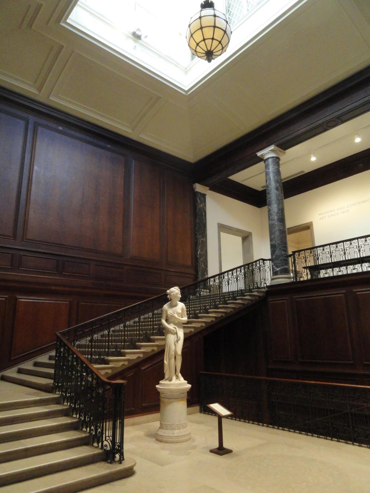 A tall skylit gallery interior with a double staircase, pale walls and framed paintings hung in a row.