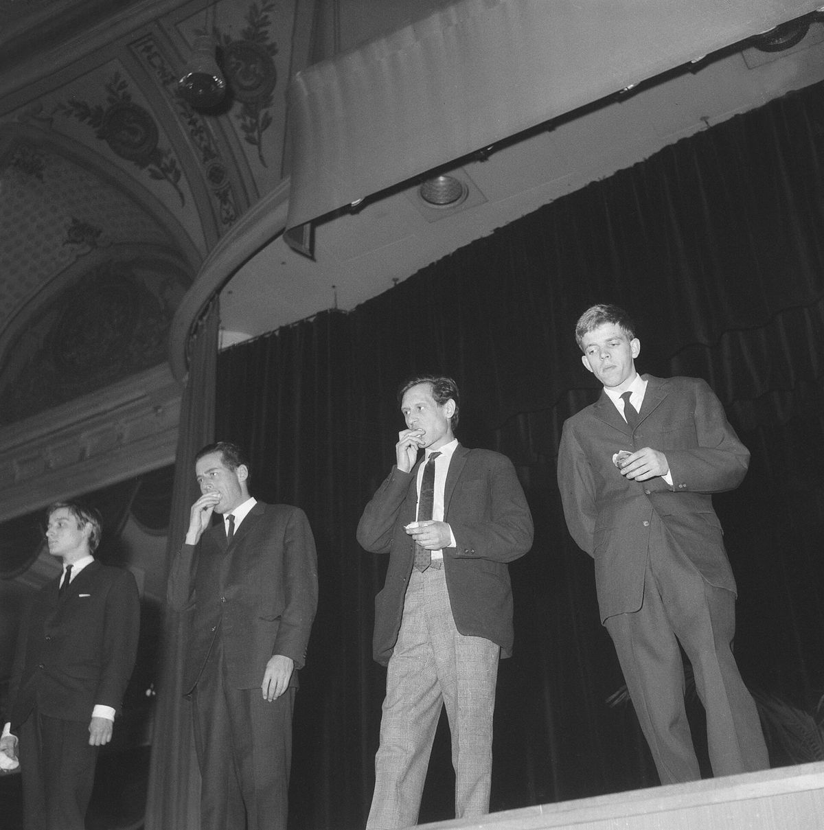 A black and white photograph from 1964 of four men in suits standing on a stage in front of a dark curtain, each raising food to his mouth.