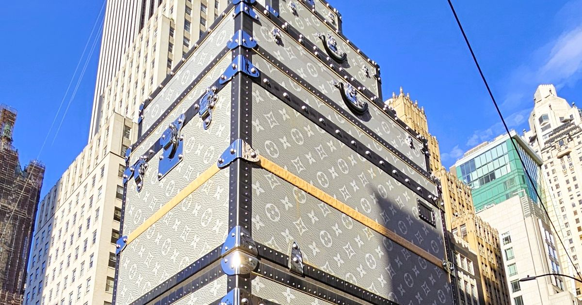 The Louis Vuitton construction shed at Fifth Avenue and East 57th Street, built to resemble a stack of monogrammed grey canvas trunks with black edging, metal corner protectors, buckles and handles, rising sixteen storeys against a clear sky.