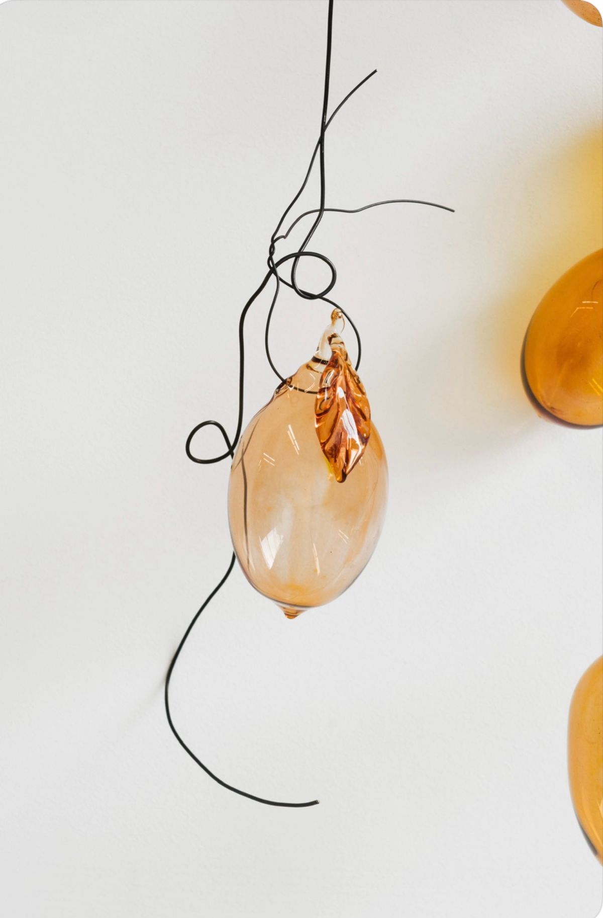 Close view of a single hand-blown glass lemon in amber, with a glass leaf, hanging from a loop of black wire against a white wall.