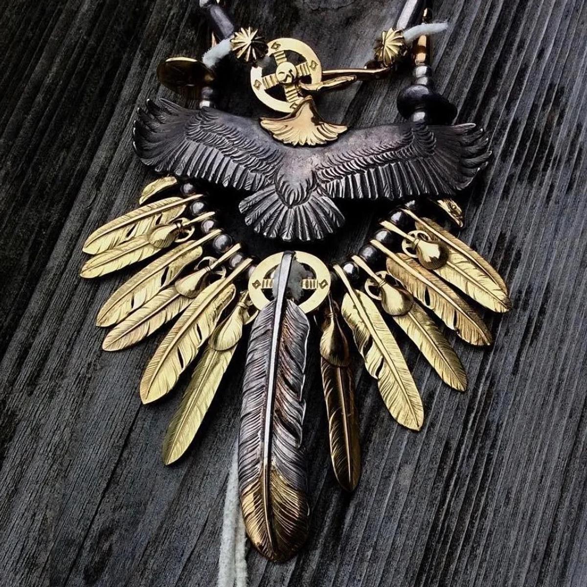 A silver pendant of an eagle with outstretched wings hung with rows of gold and silver feathers, laid on weathered grey wood.