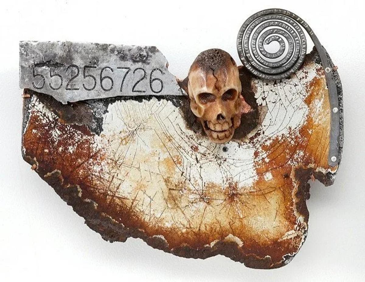 An assembled brooch of scorched wood and metal, a small carved skull at its centre beside a stamped number plate and a coiled silver spiral.