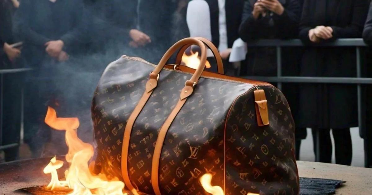 A monogrammed leather holdall sitting on a metal fire dish with flames rising around its base, a crowd standing behind a barrier watching and filming.