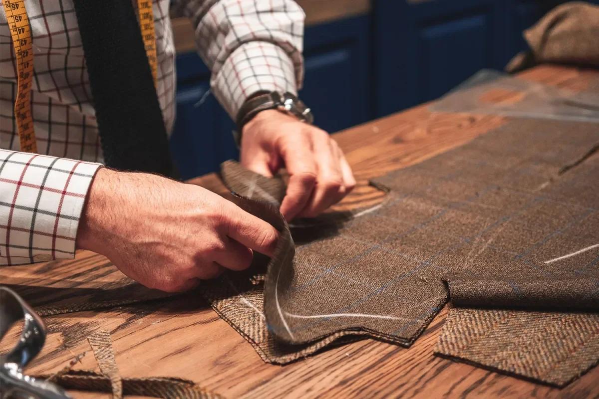 A tailor's hands smoothing a pattern piece over dark cloth on a wooden bench, a tape measure over his shoulder and shears at the edge of the table.