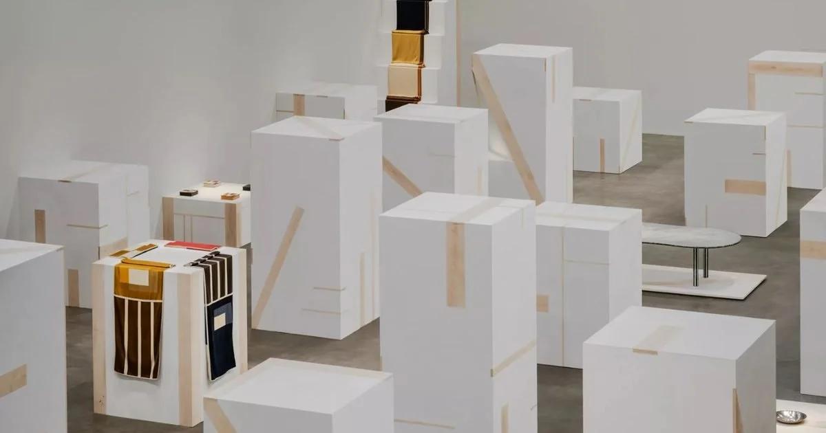 A gallery filled with white rectangular blocks arranged like a small city, one open block lined in gold and yellow, pale timber battens leaning among them.