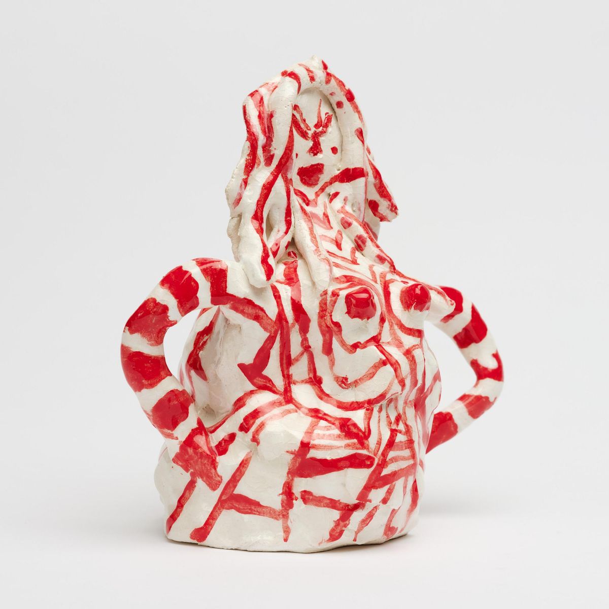 A white glazed ceramic figure with a handle, painted all over with loose red lines suggesting a face, hair and a body.