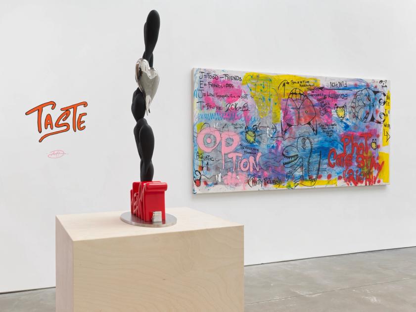 A white gallery wall with the word TASTE hand-lettered in orange and signed beneath it, a tall black and chrome sculpture of stacked forms on a pale wood plinth, and a wide painting covered in handwriting and cartoon mouths hung to the right.