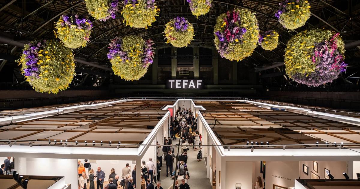 An art fair hall shot from above, dense with white booths and visitors, huge floral spheres of yellow and purple hanging under a glass roof, TEFAF lettering on the far wall.