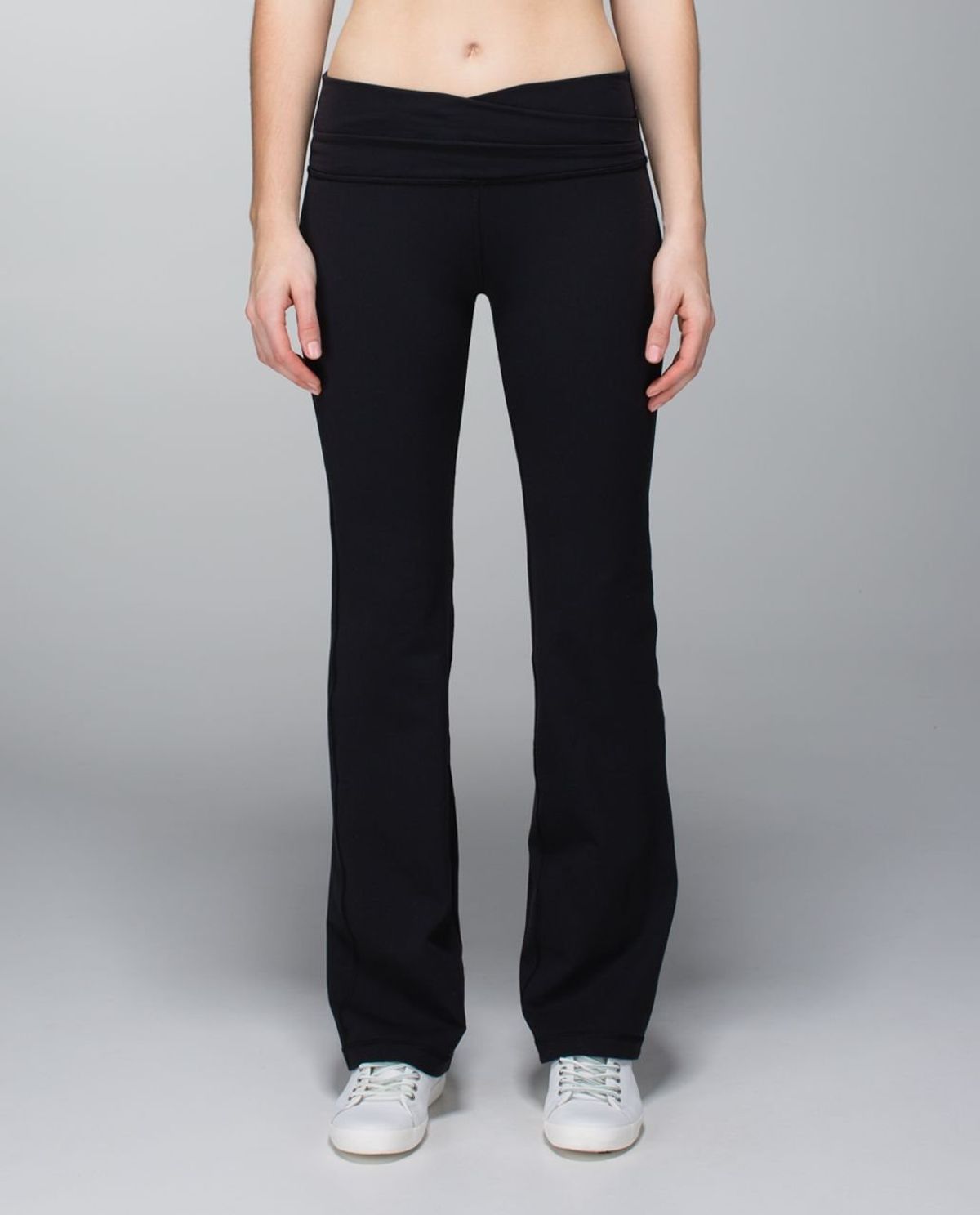 A person photographed from the waist down wearing plain black bootcut trousers with a folded waistband and white trainers, against a grey backdrop.