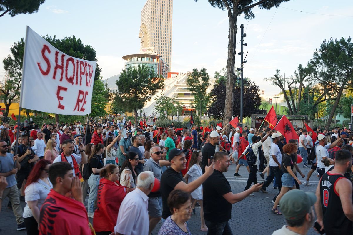 An evening march filling a boulevard in Tirana, Albanian flags carried and worn, a hand-painted banner held above the crowd, with the stepped concrete of the Pyramid of Tirana and a tower behind.