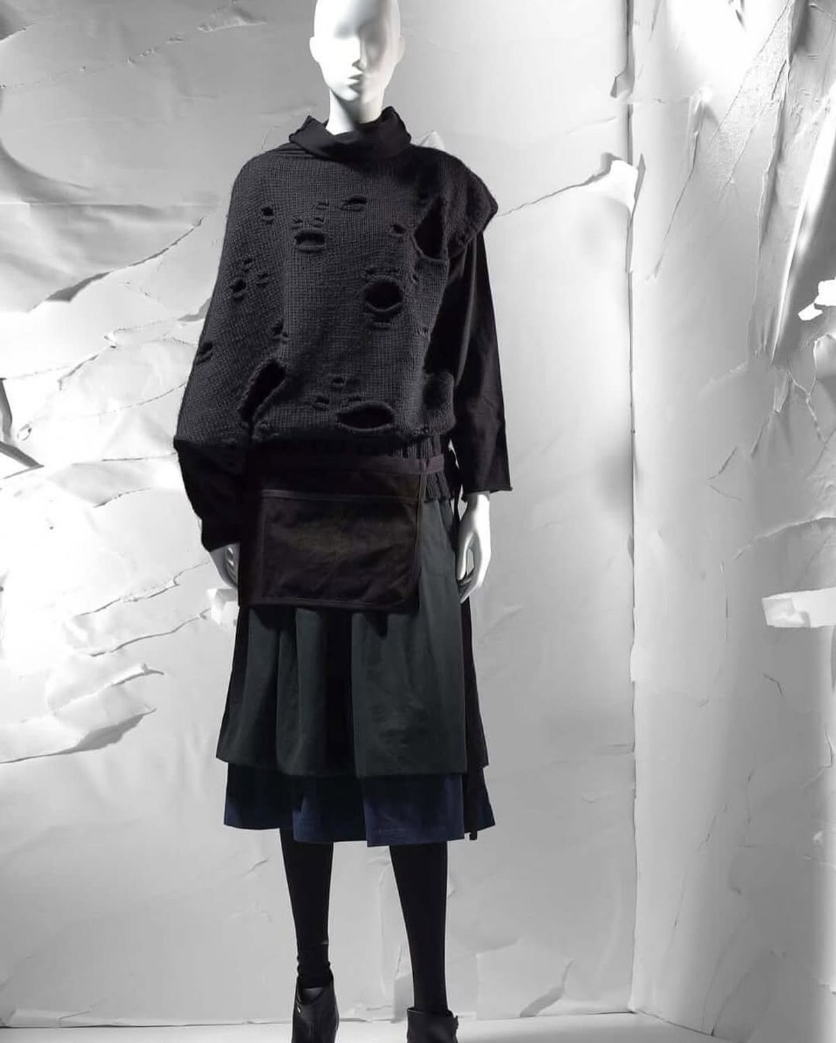A mannequin in a black holed knit sweater over a wrapped dark skirt and layered underskirt, set against a crumpled white paper backdrop.