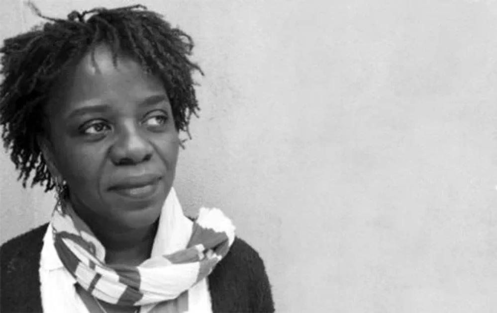 A black and white portrait of Denise Ferreira da Silva against a plain wall, wearing a checked scarf and dark cardigan, looking past the camera.