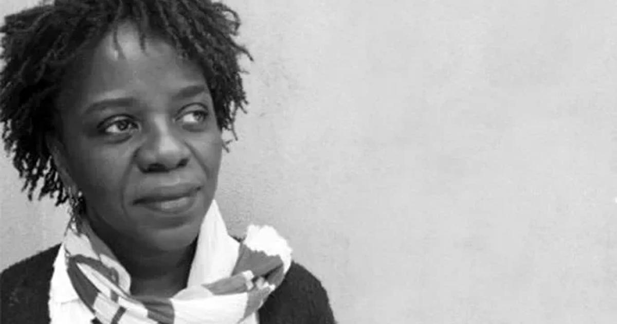 A black and white portrait of Denise Ferreira da Silva against a plain wall, wearing a checked scarf and dark cardigan, looking past the camera.