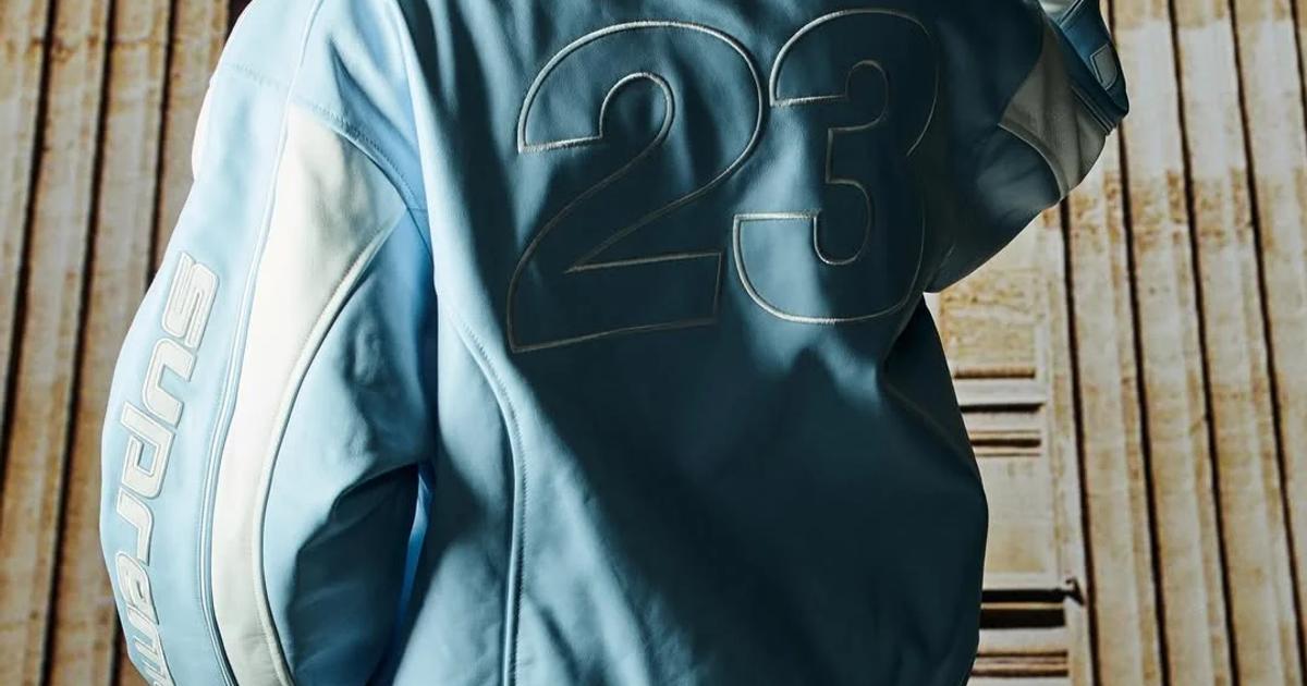 Seen from behind, a person in a pale blue hooded varsity jacket with the number 23 outlined on the back and lettering down one sleeve, standing before a stone colonnade.