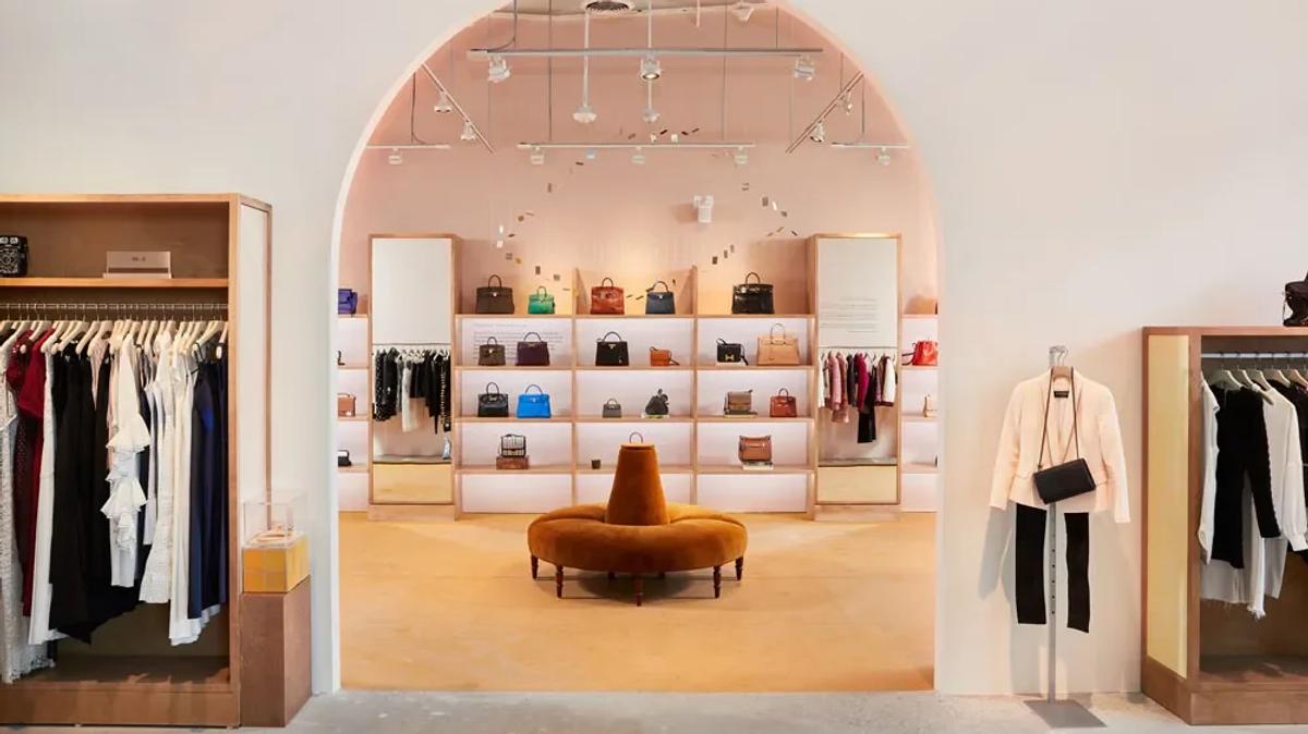 A pale pink boutique interior with an arched alcove of shelved handbags, clothing rails either side and a curved orange velvet bench at the centre.