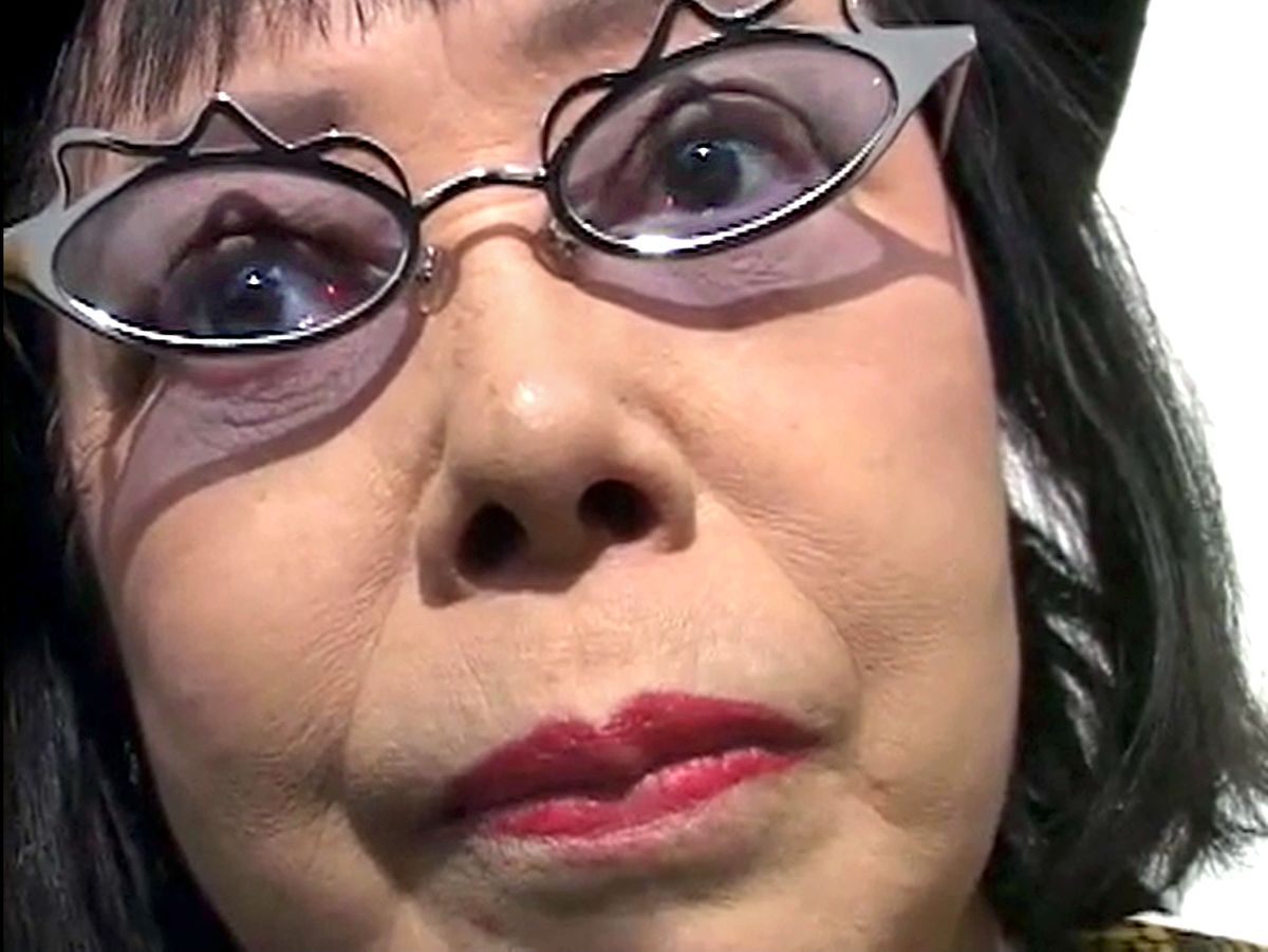 A close portrait of Yayoi Kusama, her face filling the frame, wearing distinctive dark glasses.